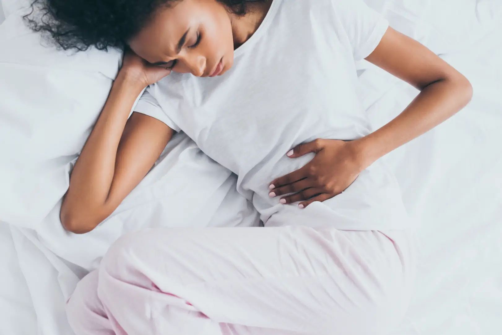 Woman lying on white bedding with one hand resting over her lower abdomen.
